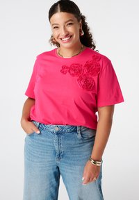 Femme souriante avec des cheveux bouclés portant un t-shirt rose avec des fleurs en tissu, un jean bleu clair, des boucles d'oreilles en or, un bracelet et un collier.