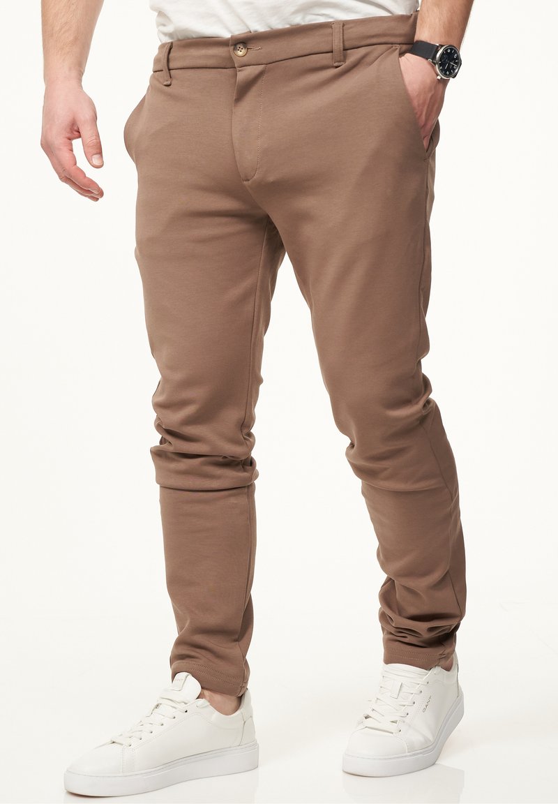 Brown slim-fit pants made from stretchy cotton blend fabric, featuring a button closure and side pockets, paired with white sneakers.