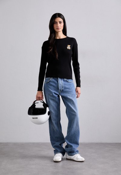 Sort ribbet langærmet sweater med et bjørneapplikation, lyseblå løse jeans og hvide sneakers, mens der holdes en hvid motorcykelhjelm.