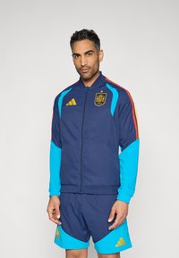 Chaqueta deportiva azul marino con mangas azules y detalles rojos, que presenta el emblema nacional de España y el logo dorado de Adidas. Acompañada de pantalones cortos a juego.