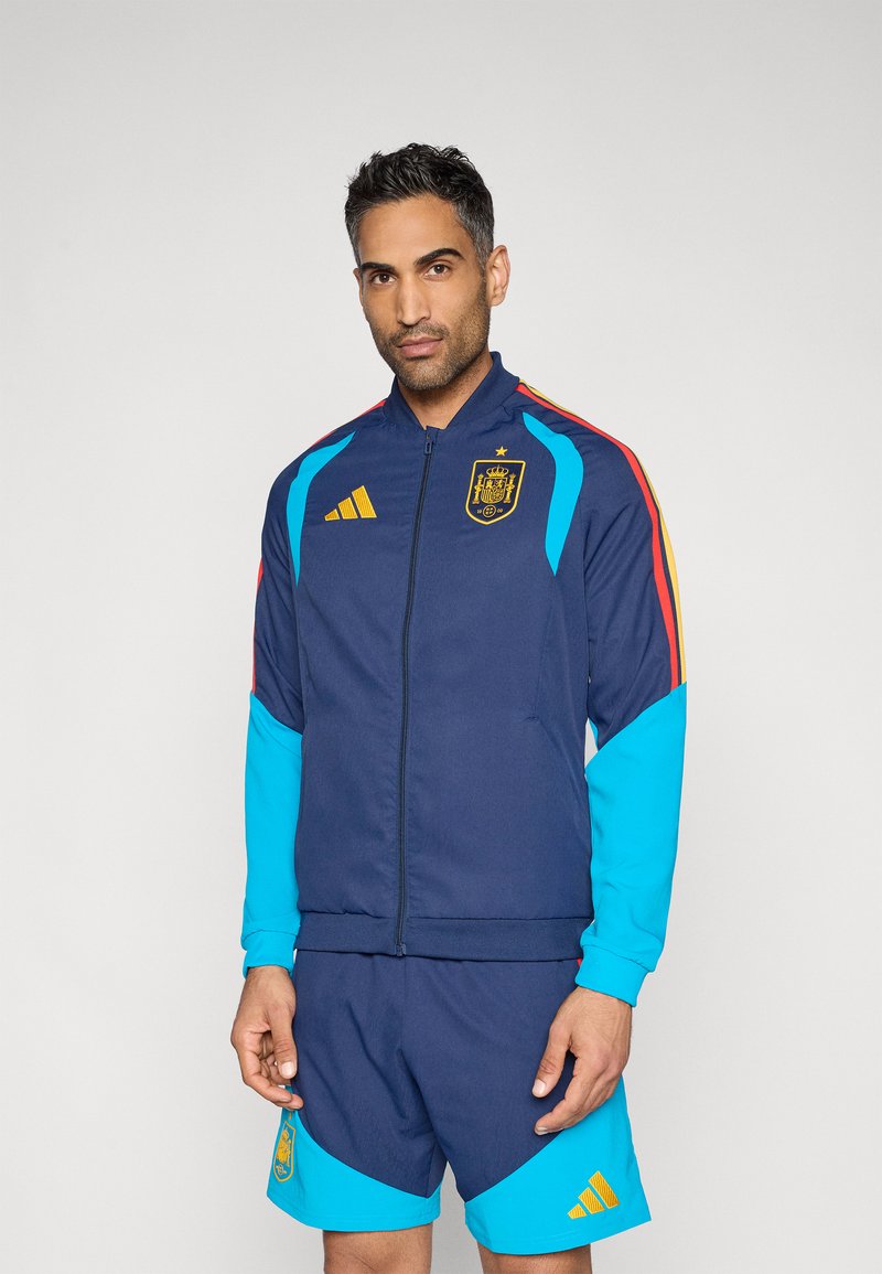 Chaqueta deportiva azul marino con mangas azules y detalles rojos, que presenta el emblema nacional de España y el logo dorado de Adidas. Acompañada de pantalones cortos a juego.