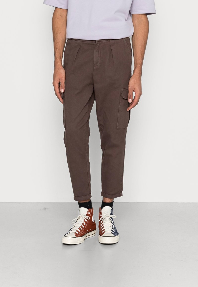 Brązowe spodnie zwężane z plisami, z boczną kieszenią typu cargo. Do tego dwukolorowe trampki za kostkę, jedna czerwona, druga niebieska.