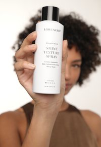 Flacon vaporisateur cylindrique blanc avec un capuchon noir, étiqueté "LÖWENGRIP Bounce Back Shine Texture Spray," 200 mL, finition texturée, impression claire.
