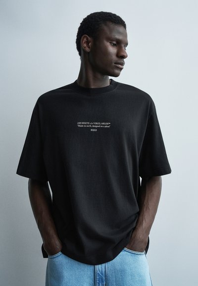 Homme portant un t-shirt Off-White noir avec le texte « Made on earth, designed on a plane » et un jean bleu clair, mains dans les poches, regardant vers la droite.