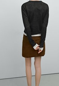 Schwarzer Strickpullover, der über einem hellen Langarmoberteil getragen wird, kombiniert mit einem braunen Wildlederrock. Details umfassen lange Ärmel und ein tailliertes Design.