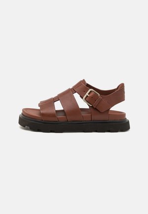 Sandal in pelle marrone con ampie cinghie, fibbia regolabile e suola spessa in gomma nera, mostrato in profilo laterale su sfondo bianco.