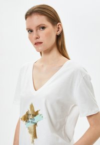 Camiseta de algodón blanca con escote en V y mangas cortas. Presenta un estampado gráfico en dorado y azul claro con texto en el centro.