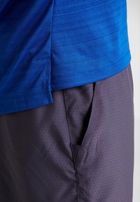 Blau strukturiertes Sportshirt kombiniert mit grauer Shorts, die eine sichtbare Seitentasche und ein perforiertes Muster zur Belüftung aufweist.