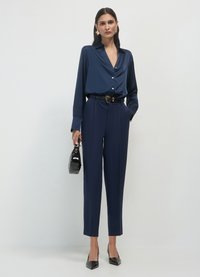 Navy-satines Hemd mit langen Ärmeln und V-Ausschnitt, kombiniert mit hochgeschnittenen, maßgeschneiderten Hosen und einem schwarzen Gürtel. Die Accessoires umfassen eine kleine schwarze Handtasche.