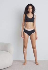 Zwarte naadloze bralette en bijpassende bikini, beide met een gladde textuur. Model staat op een licht tapijt met een ronde beige fauteuil in de buurt.