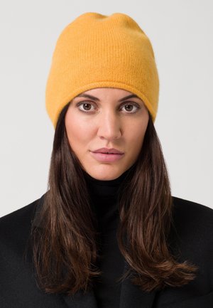 Geel wollen beanie met een ontspannen pasvorm, voorzien van een naadloze bovenkant en textuurbreisels. Draag het met een zwarte coltrui en jas.