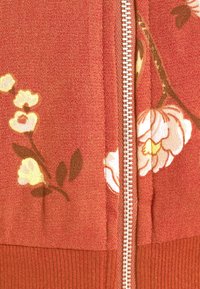Veste zippée rouge à motifs floraux, en tissu texturé, avec des motifs floraux crème et jaune, et un ourlet et des poignets côtelés assortis.