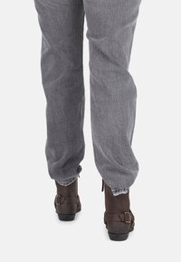 Pantalones de pana gris con un acabado texturizado, que presentan un corte relajado y un largo hasta el tobillo, combinados con botas de cuero marrón con correa de hebilla.