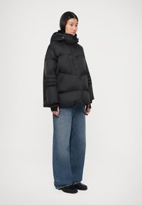 Jachetă puffer neagră cu glugă, având un design pătrat, textură matlasată, buzunar frontal și manșete elastice. Asortată cu blugi din denim cu picioare largi.