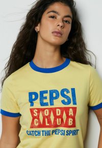 Geel T-shirt met blauwe randen aan de kraag, met de tekst "PEPSI SODA CLUB" in blauwe en rode grafische letters en de slogan "CATCH THE PEPSI SPIRIT".