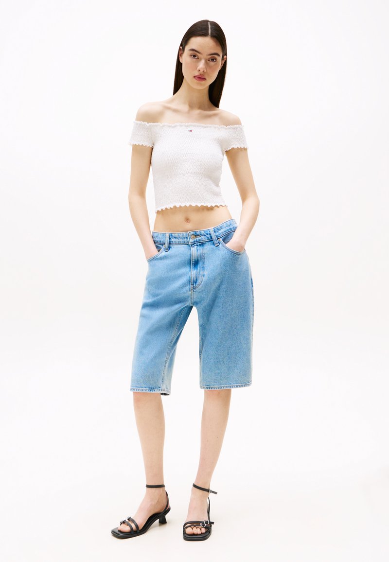 Fehér, vállatlan, textúrált kialakítású crop top, világoskék farmer berber nadrággal párosítva, amely zsebekkel és laza fit állással rendelkezik. Fekete, magas sarkú szandál.