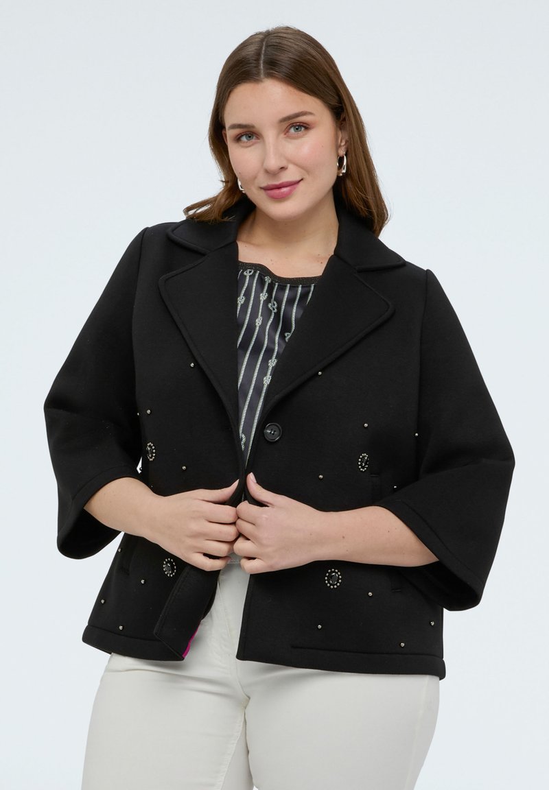 Mujer con un abrigo corto negro de mangas anchas y tachuelas decorativas sobre una blusa a rayas y pantalones blancos, de pie contra un fondo liso.