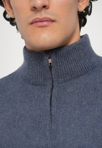 Άνδρας που φοράει μια μπλε υψωμένη πλεκτή μπλούζα με έναν ημιτελή μαύρο φερμουάρ στον μπροστινό γιακά.