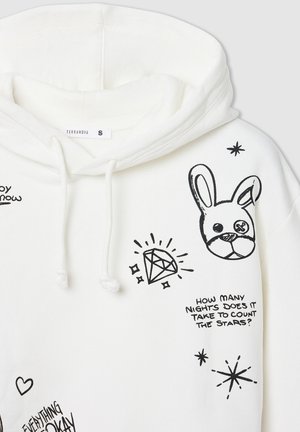 Biała bluza z kapturem posiada czarne graficzne wzory, w tym króliczka, diament i tekst. Sznurki przy kapturze mają miękką teksturę.