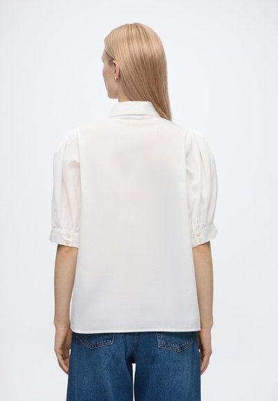 Femme aux cheveux blonds lisses portant un chemisier blanc à manches courtes bouffantes et un jean en denim bleu, vue de dos sur fond blanc.