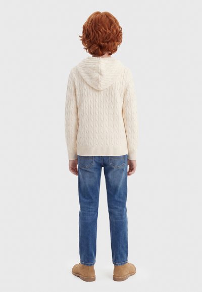 Παιδί με κόκκινα σγουρά μαλλιά που φοράει μία κρεμ πλεκτή μπλούζα με κουκούλα, μπλε τζιν και ταμπα σουέτ μπότες, στέκεται γυρισμένο προς τα πίσω σε λευκό φόντο.