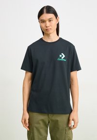Camiseta de algodón negra con cuello redondo, que presenta un logo gráfico verde y un diseño de estrella en el lado izquierdo del pecho. Mangas rectas.