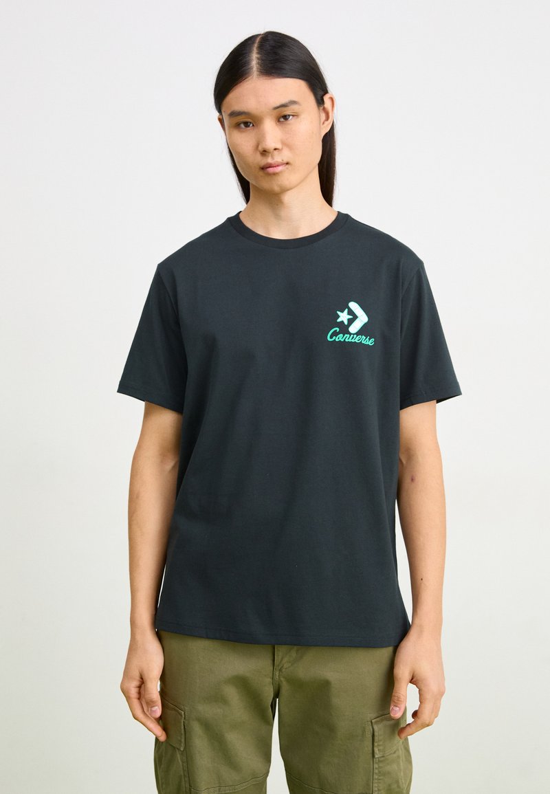 Camiseta de algodón negra con cuello redondo, que presenta un logo gráfico verde y un diseño de estrella en el lado izquierdo del pecho. Mangas rectas.