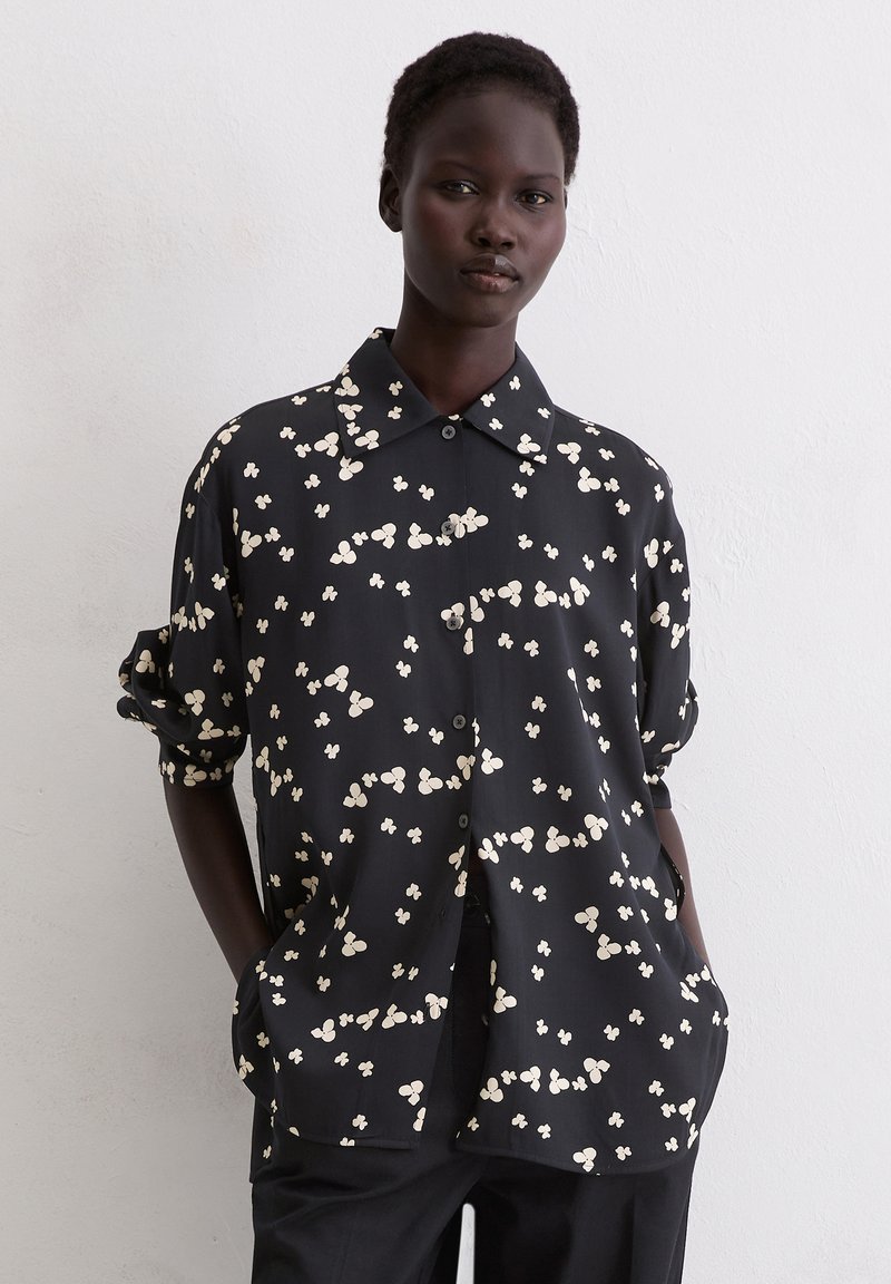 Zwarte overhemden met knopen, gemaakt van gladde stof, met een wit bloemenpatroon. Het overhemd heeft een kraag en opgerolde mouwen.