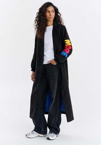 Kobieta z długimi kręconymi włosami, ubrana w długi czarny płaszcz z kolorowymi literami na rękawie, białą koszulę, szerokie czarne dżinsy i białe trampki.