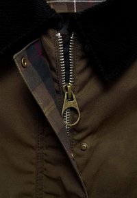 Nærbillede af en brun jakke lynlås med et stort messingtræk, sort corduroy-krave og ternet stofforing synlig bag lynlåsen.