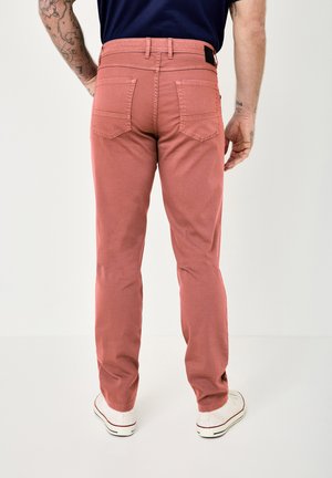 Mann trägt staubrosa gerade geschnittene Hose, weiße Schuhe und ein marineblaues Hemd, steht mit dem Rücken zur Kamera vor einem einfarbigen Hintergrund.