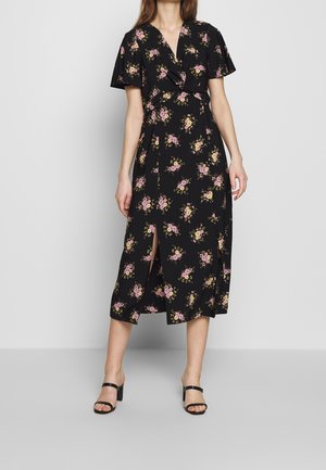 Femme portant une robe midi noire à imprimé floral rose et jaune, manches courtes flottantes, col en V croisé, fente devant, et sandales à talons noires.