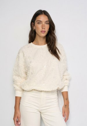 Mujer con cabello largo y oscuro, vestida con una camiseta blanca texturizada de manga larga y pantalones blancos, de pie contra un fondo liso y claro.