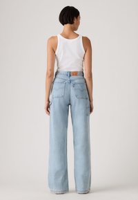 Γυναίκα με κοντά σκούρα μαλλιά που φοράει άσπρο αμάνικο τοπ και ανοιχτό μπλε τζιν με ψηλή μέση και φαρδιά μπατζάκια, όρθια με την πλάτη προς τα έξω.