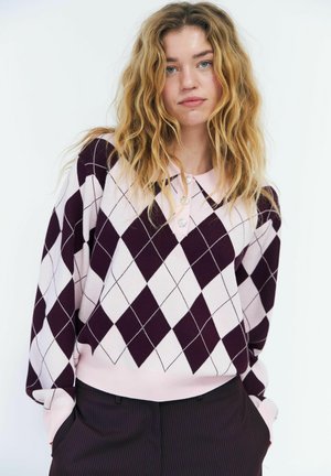 Giovane donna con capelli biondi mossi che indossa un maglione rosa e viola scuro a rombi e pantaloni scuri, in piedi con le mani nelle tasche su sfondo bianco.