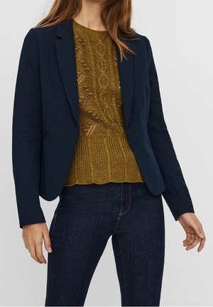 Frau trägt einen dunkelblauen Blazer über einem senffarbenen Zopfpullover und dunkelblaue Jeans vor einfarbigem Hintergrund.