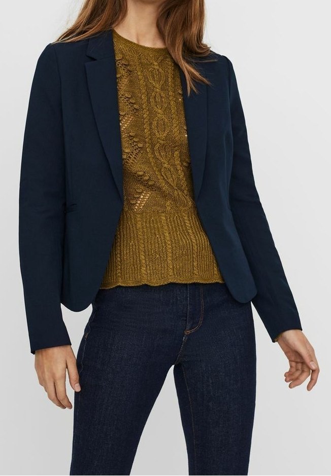 Femme portant un blazer bleu marine foncé sur un pull en maille torsadée moutarde et un jean bleu foncé sur un fond uni.