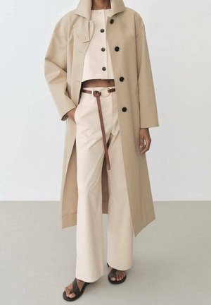Persoon draagt een beige trenchcoat over een crèmekleurige, knoopje-erop cropped top en een broek met hoge taille en bruine riem, staat met één hand in de zak en zwarte sandalen.