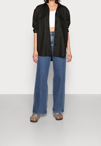 Black oversized shirt with two front pockets, worn over a white crop top. Paired with high-waisted blue wide-leg jeans and gray sandals.