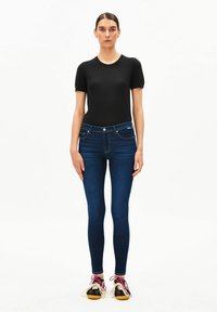 Frau trägt ein schwarzes kurzärmeliges Oberteil, dunkelblaue Skinny-Jeans und schwarze Sneakers mit pinken und weißen Schnürsenkeln, steht vor einem schlichten weißen Hintergrund.