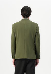 Man with dark hair wearing an olive green blazer and black pants, standing facing a plain white background.