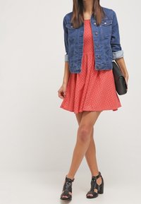 Robe texturée corail avec coupe évasée, veste en denim bleu, sac à main noir et escarpins noirs ouverts à talons épais avec brides à la cheville.