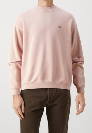 Person wearing a light pink crewneck sweatshirt with ribbed cuffs and hem, paired with dark brown pants, shown from neck to hips.