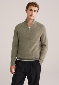 Sweter robiony na drutach w stonowanym oliwkowym zielonym kolorze, z kołnierzem na ćwierć zamka i prążkowaną teksturą; noszony na białej koszulce i w zestawie z czarnymi spodniami.