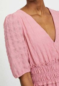 Rosa, texturerad klänning med djup V-ringning, korta puffärmar och samlad midja. Materialet har ett subtilt rutmönster.