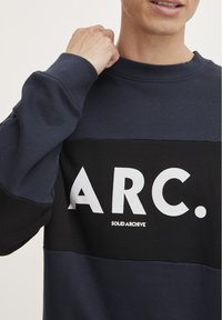 Persona che indossa un felpa blu navy e nera con una grande scritta bianca "ARC." e un testo più piccolo "SOLID ARCHIVE", sollevando un pugno vicino al collo.