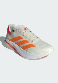 Zapatilla deportiva de un tono beige claro con tres rayas naranjas, parte superior texturizada y suela blanca acentuada con rojo. Presenta un collar acolchado y cordones.