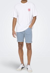 Valkoinen lyhythihainen t-paita, jossa on vaaleanpunainen grafiikka rinnassa, yhdistettynä vaaleansinisiin käärityn lahkeen farkkushortseihin ja valkoisiin tennareihin, joissa on mustat yksityiskohdat.