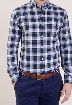 Homme portant une chemise à manches longues à carreaux verts, blancs et bleu marine, rentrée dans un pantalon bleu marine avec une ceinture en cuir marron.