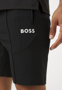 Șorturi atletice negre, realizate dintr-un material texturat, având un buzunar lateral și un logo alb "BOSS" proeminent pe coapsă.